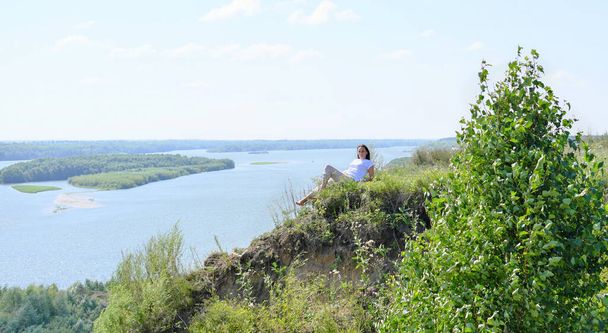 панорамний вид на високий берег річки і жінку, що сидить на блакитному фоні. красива жінка насолоджується перебуванням на березі річки. час для внутрішнього спокою, релаксації та медитації на відкритому повітрі
.  - Фото, зображення