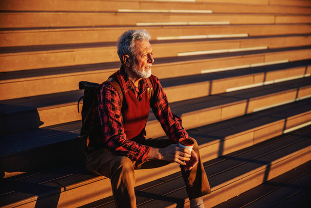 Homme âgé barbu assis dans les escaliers à l'extérieur, regardant quelque chose et buvant du café pour aller. - Photo, image