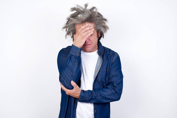 Portrait intérieur d'un homme triste avec une coupe de cheveux élégante, portant des vêtements décontractés, faisant un geste de facepalm tout en se tenant sur fond blanc - Photo, image