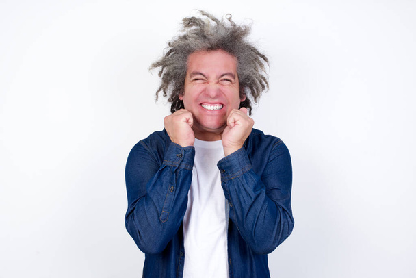 Porträt eines Mannes mit afrograuen Haaren, der mit geschlossenen Augen und Händen vor dem Gesicht Begeisterung und Glück ausdrückt, während er auf weißem Hintergrund breit lächelt.  - Foto, Bild
