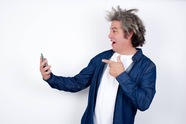 Портрет счастливого веселого человека с седыми волосами на белой стене, получающего хорошие новости по электронной почте и празднующего успех, стоя и глядя на мобильный телефон - Фото, изображение