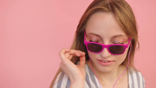 Fiatal fehér nő napszemüvegben fülhallgatóval hallgatja a zenét. Rózsaszín háttérrel, közelről - Fotó, kép