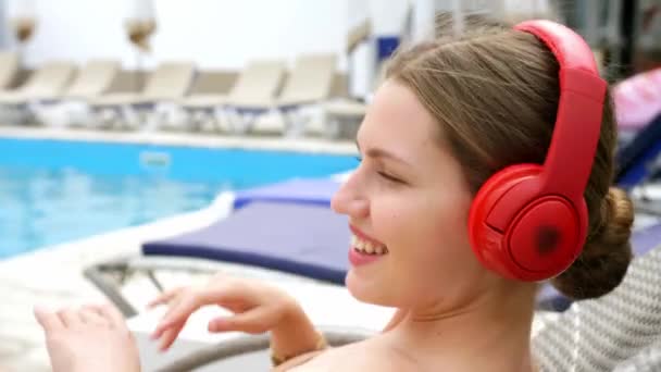Mooie jonge vrouw luisteren naar muziek in de buurt van zwembad in resort - Video
