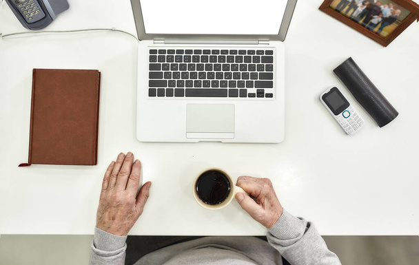 Top view of aged male employee senior intern holding a cup of coffee while sitting at desk in modern office, Notebook, picture frame, cell phone, glasses and laptop on the table - Photo, Image