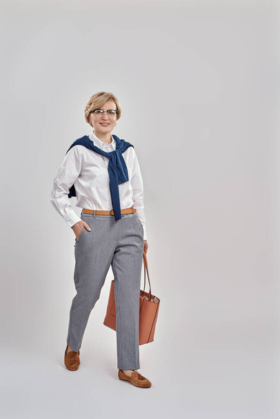 Full length shot of elegant middle aged caucasian woman wearing business attire and glasses smiling at camera while posing, standing isolated over grey background - Фото, зображення