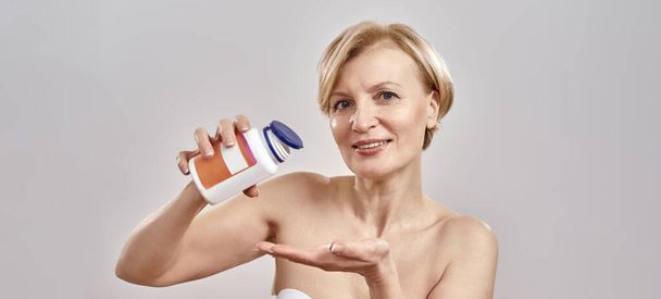 Portrait of middle aged caucasian woman pouring the pills out of the bottle and smiling at camera while standing isolated over grey background. Healthcare concept - Photo, Image