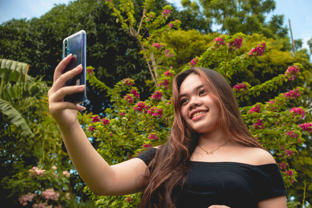 Una joven y de moda chica del sudeste asiático se toma una selfie de sí misma contra un entorno de jardín. Usar una blusa negra sin hombro y sostener un teléfono inteligente azul. - Foto, imagen