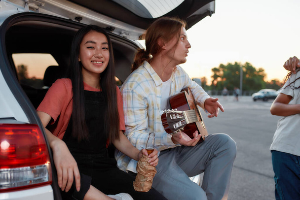 Two young casually dressed friends of different nationalities sitting in an opened car trunk outside on a parking site smiling and playing guitar - Foto, immagini