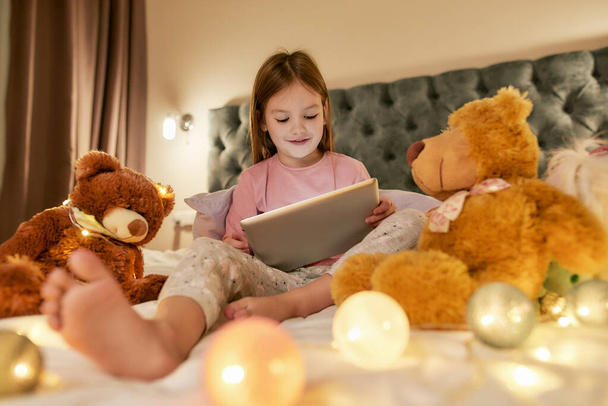 A cute little girl wearing pyjamas sitting alone on a huge bed barefoot enjoying her time watching cartoons on a tablet and smiling before going to sleep with her fluffy toys and garlands - Zdjęcie, obraz