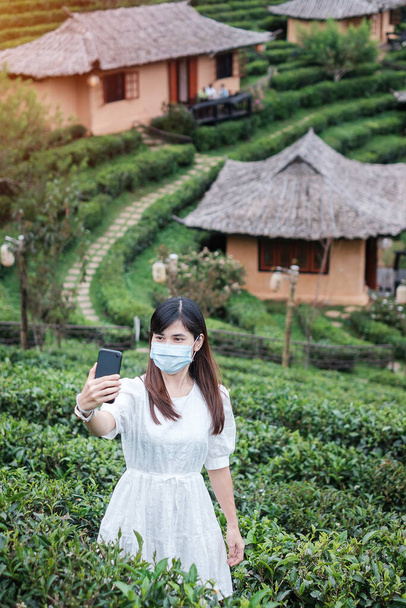 Mujer turista feliz con máscara facial quirúrgica y el uso de teléfono inteligente móvil en el hermoso jardín de té, protección contra la enfermedad por Coronavirus (Covid-19) infección. Nuevo Normal, seguridad de viaje, vacaciones y vacaciones - Foto, imagen