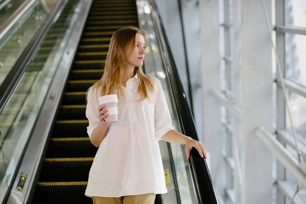 retrato de la hermosa mujer joven en camisa blanca con café cerca de escaleras mecánicas  - Foto, Imagen