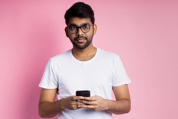 Portrait of happy indian young man in glasses, holding cell phone, sending text message, reading good news, posing against pink background. Technology, communication. - Foto, imagen