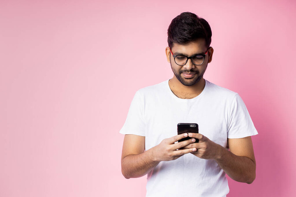 Indian man holding a smartphone in his hands, wearing glasses, white t-shirt, looking at the phone on a pink background - Foto, immagini