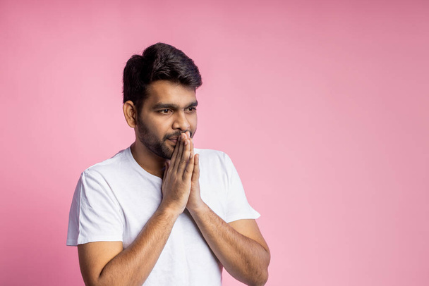 Closeup of young indian male in white t shirt, praying with palms clasped together, asking the God for good luck, success, health, on pink background with copy space. Human emotion, belief concept. - Photo, Image