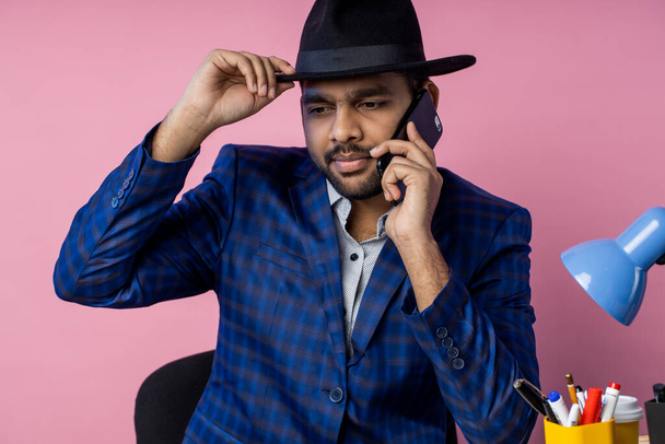 bearded stylish young businessman in formal wear, having phone conversation with partner, discussing terms of deal, drinking coffee, siiting at table in office, against pink wall. - Foto, Bild