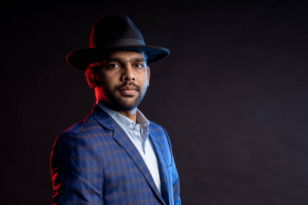 handsome confident bearded Indian man, businessman wearing shirt, checked suit, black hat, looking aside, standing isolated on dark background. - Фото, зображення