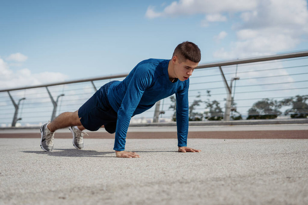 Morning exercise and workout concept. Low angle view of adult strong and focused sportsman making sport training outdoors alone, doing push ups exercise and standing in plank pose - Photo, Image