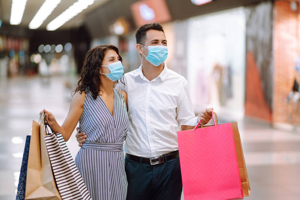 Beautiful young loving couple  in protective sterile medical mask carrying shopping bags and enjoying together. Shopping in the coronavirus epidemic. Purchases, black friday, discounts, sale concept. - Foto, Bild