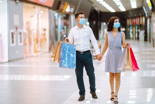 Retrato de una pareja con bolsas de compras en máscara médica estéril protectora en el centro comercial. Compras en la pandemia del coronavirus. Gente, venta, amor, viernes negro y concepto de felicidad. - Foto, imagen