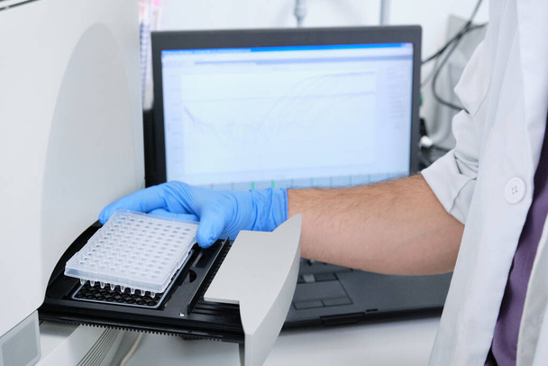 Ερευνητής τοποθετεί την πλάκα PCR στον θερμικό κυκλωτή για ενίσχυση του DNA. Καμπύλη χάρτη στην οθόνη πίσω. Δοκιμή PCR του Coronavirus. - Φωτογραφία, εικόνα