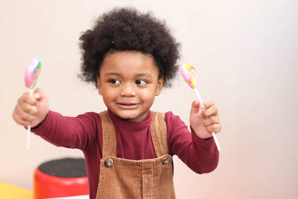 Retrato de adorable niño afroamericano con pelo rizado sosteniendo dulces dulces, alegre niño disfrutar con colorido arco iris piruleta - Foto, Imagen
