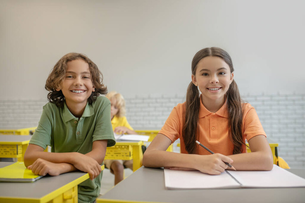 Two schoolchildren sitting at the desk and looking happy - Fotó, kép