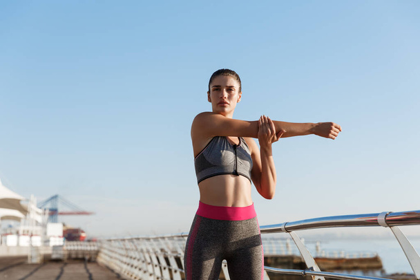 ジョギングトレーニングの前に彼女の体を伸ばすフィットネス服の美しい健康的な女性のイメージ。海辺の遊歩道に立つ女性ランナー、海の近くでのトレーニング - 写真・画像