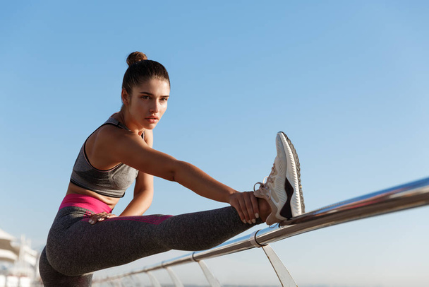 Εικόνα από ελκυστική τακτοποίηση γυμναστικής κορίτσι προπόνηση σε μια προβλήτα. Αθλητής που τεντώνει τα πόδια και προθέρμανση πριν από την προπόνηση τζόκινγκ στον παραλιακό πεζόδρομο - Φωτογραφία, εικόνα