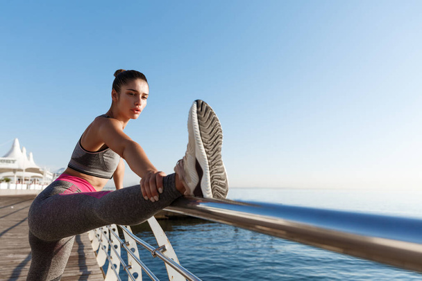 Outdoor shot van aantrekkelijke gezonde vrouw in sportkleding, strekken haar benen en warming-up voor joggen workout in de buurt van zee - Foto, afbeelding