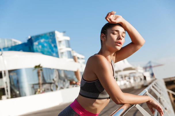 Close-up of attractive young fitness woman leaning on handrail, wiping sweat off forehead after workout, finish jogging on the seaside promenade - Fotó, kép