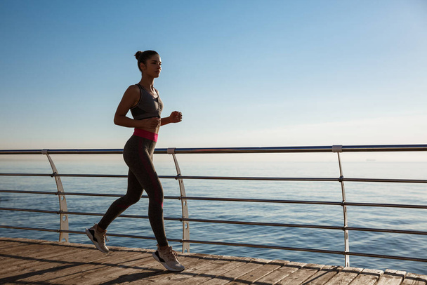 Oldalnézet fitness nő fut végig tengerparti sétányon, edzés a friss levegőn, közel a tengerhez - Fotó, kép