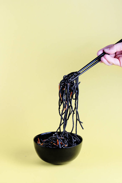 Black wok noodles with sesame seeds and carrots in a black cup, hand takes a bundle of noodles with black chopsticks on a light yellow background - Photo, Image