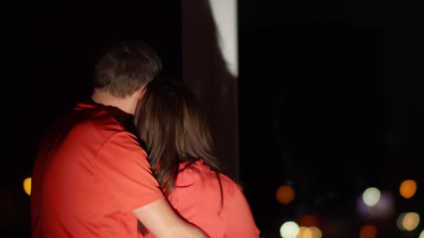 Een liefdevol stel staat 's nachts aan het raam te knuffelen, achteraanzicht - Video