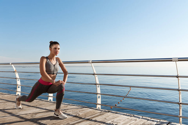 Foto al aire libre de la sonriente mujer fitness estirándose y ejercitándose en el paseo marítimo - Foto, imagen