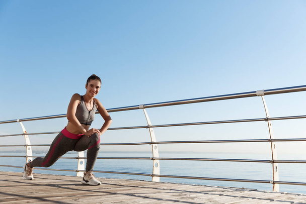 Foto al aire libre de atractiva y saludable entrenamiento de mujer fitness cerca del mar, estirando las piernas antes de trotar - Foto, Imagen