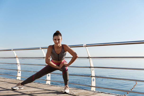 Νεαρή ελκυστική αθλήτρια προπόνηση κοντά στη θάλασσα. Γυμναστική γυναίκα τεντώνει τα πόδια της πριν από το τρέξιμο και την κατάρτιση στην παραλία - Φωτογραφία, εικόνα