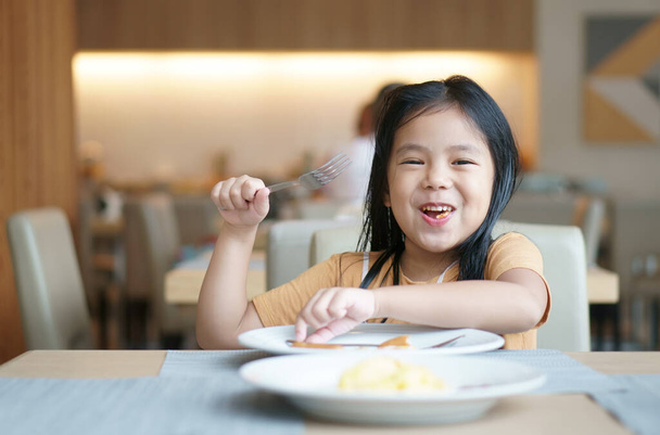 アジアの子供のかわいいまたは子供の女の子は、白い料理とテーブルの上で空腹のためにおいしい食べ物を食べることを楽しむためにフォークを保持笑顔ランチや朝食とレストランやフードコートで朝の食欲をそそる - 写真・画像