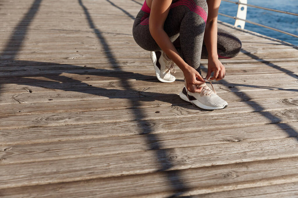 Recortado tiro de mujer fitness atándose los cordones de los zapatos, prepararse para trotar en el paseo marítimo - Foto, imagen