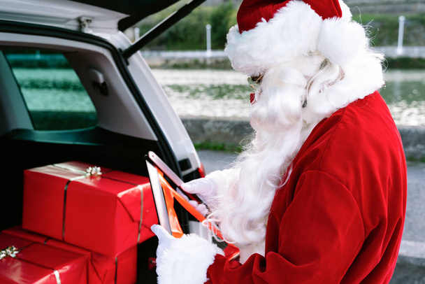Joulupukin muotokuva auton takakontti auki täynnä lahjoja ottaen hätäkolmion ulos autosta, koska hänen autonsa on hajonnut ja hänellä on kasvonaamio koronaviruksen kovettumisen vuoksi19 pandemia jouluna 2020 - Valokuva, kuva