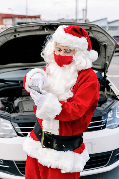 Joulupukin muotokuva kirjoittaa jotain matkapuhelimeensa huolissaan ja autonsa huppu auki, koska hänen autonsa on hajonnut eikä hän voi toimittaa lasten lahjoja jouluna 2020 - Valokuva, kuva