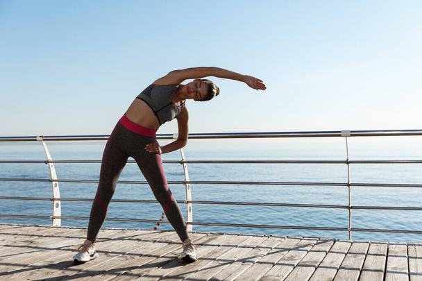 Outdoor shot of attractive and fit sportswoman workout on a pier. Girl stretching and training alone at the seaside - Photo, Image