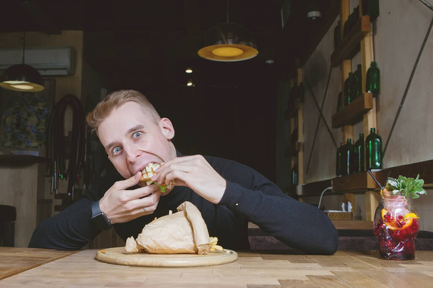 Young blond guy enjoys eating a sandwich at a table in a cafe. - Фото, изображение