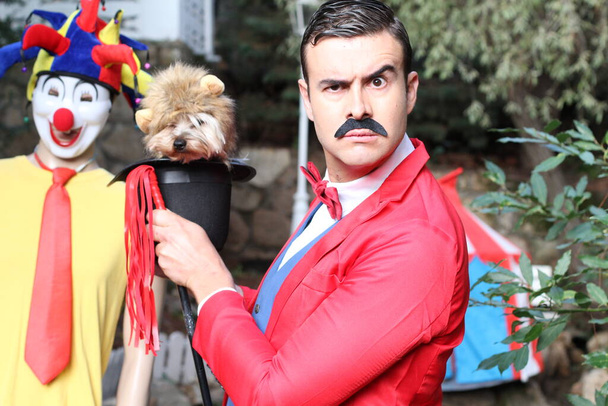 retrato de guapo joven mago en traje rojo sosteniendo lindo cachorro en circo - Foto, Imagen