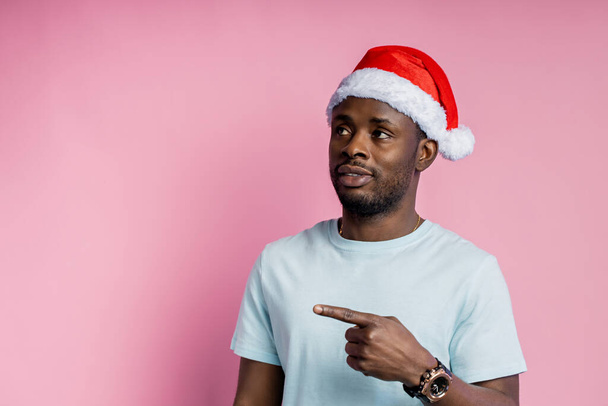 Handsome serious bearded african american young man wearing red Santa hat, white t shirt, looking at camera with calm, confident expressions, isolated over pink background. Christmas concept. - Foto, imagen