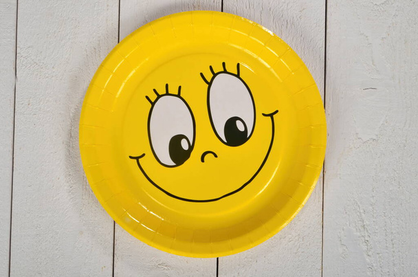 Χαρούμενο χαμογελαστό πρόσωπο σε κίτρινο χάρτινο πιάτο. Άσπρο ξύλινο φόντο. Χαρούμενο κίτρινο emoticon χαμογελώντας - Φωτογραφία, εικόνα