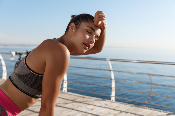 Beeld van mooie jonge fitnessvrouw hijgend en vegen zweet van het voorhoofd na joggen, finish run of workout, staande op een pier - Foto, afbeelding