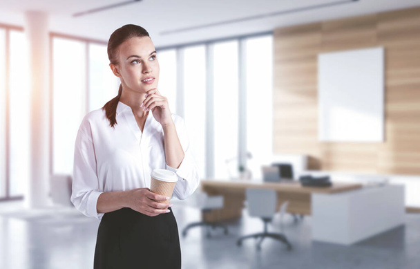 Portrait of pensive young businesswoman holding paper cup of coffee in blurry office. Concept of brainstorming - Fotó, kép