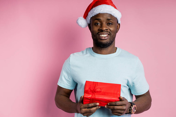 Komea onnellinen afrikkalainen amerikkalainen mies yllään Santa hattu, tilalla punainen lahjapakkaus, hymyillen, antaa lahja tyttöystävä, eristetty yli vaaleanpunainen tausta. Joulun käsite. - Valokuva, kuva