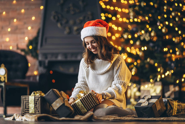 Christmas, New Year. Pretty woman in warm sweater, socks and Christmas hat, sitting on the floor at home with gift boxes and new year tree and lights behind - 写真・画像