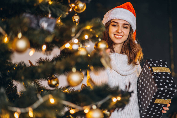 Christmas, New Year. Pretty woman in warm sweater, socks and Christmas hat, decorating new year tree and holding gif box - Фото, изображение